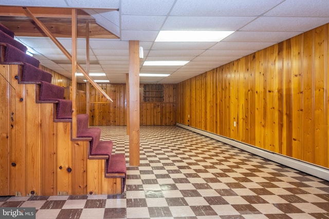 basement with tile patterned flooring, a baseboard heating unit, wooden walls, and a paneled ceiling