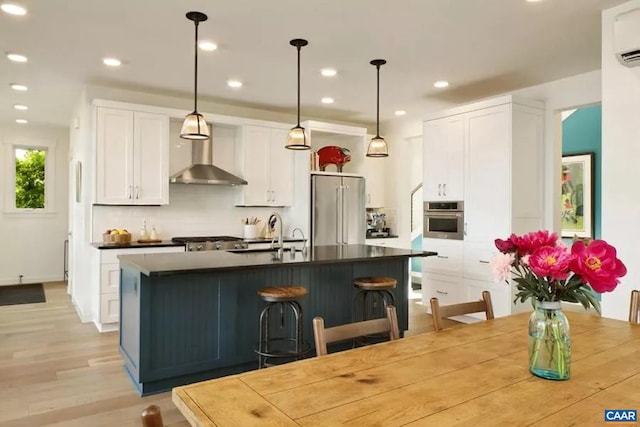 kitchen with a kitchen island with sink, white cabinets, wall chimney exhaust hood, decorative light fixtures, and stainless steel appliances