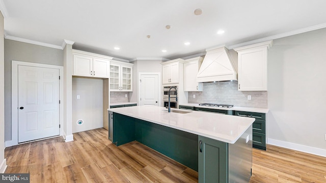 kitchen featuring a kitchen island with sink, stainless steel gas cooktop, light countertops, glass insert cabinets, and custom range hood