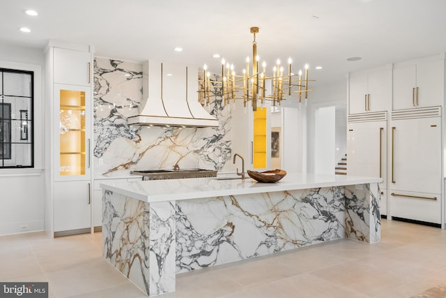 kitchen with decorative light fixtures, premium range hood, paneled fridge, an island with sink, and white cabinets