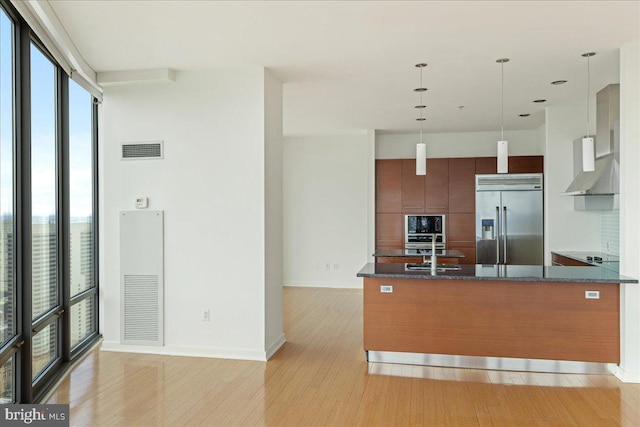 kitchen with wall chimney range hood, hanging light fixtures, built in appliances, dark stone countertops, and a wall of windows