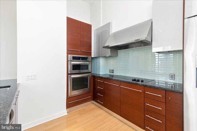 kitchen with wall chimney exhaust hood, stainless steel double oven, backsplash, black electric cooktop, and light wood-type flooring