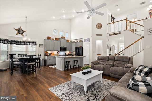 living room with ceiling fan with notable chandelier, a high ceiling, sink, and dark wood-type flooring