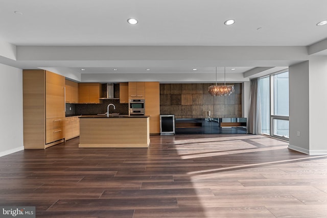 kitchen with light brown cabinets, an inviting chandelier, a center island with sink, wall chimney range hood, and decorative light fixtures