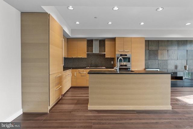 kitchen with gas cooktop, dark wood-type flooring, wall chimney range hood, light brown cabinets, and an island with sink