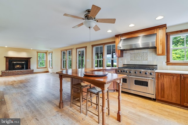 kitchen featuring backsplash, wall chimney range hood, range with two ovens, a fireplace, and light hardwood / wood-style floors