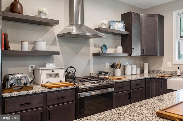 kitchen with wall chimney exhaust hood, stainless steel gas range oven, light stone counters, and dark brown cabinetry