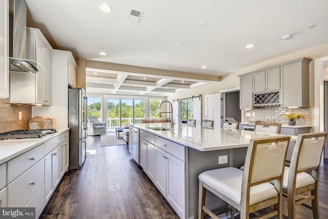 kitchen featuring a center island with sink, stainless steel appliances, a barn door, coffered ceiling, and wall chimney exhaust hood