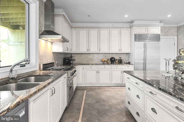 kitchen featuring wall chimney exhaust hood, high end appliances, sink, and dark stone counters