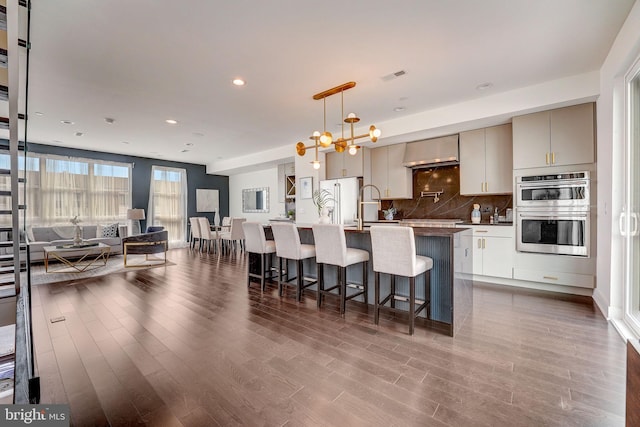 kitchen featuring appliances with stainless steel finishes, backsplash, exhaust hood, pendant lighting, and a center island with sink