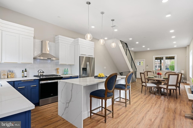 kitchen featuring appliances with stainless steel finishes, wall chimney exhaust hood, blue cabinets, a center island, and hanging light fixtures