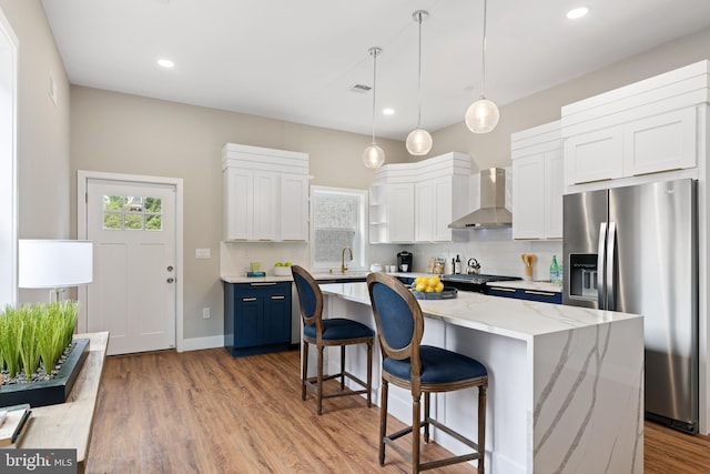 kitchen with wall chimney range hood, a kitchen island, stainless steel refrigerator with ice dispenser, blue cabinets, and white cabinets