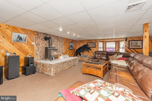 living room featuring a drop ceiling, a wood stove, carpet, and wooden walls