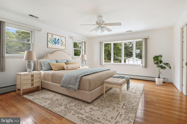 bedroom featuring ceiling fan, light hardwood / wood-style floors, multiple windows, and a baseboard heating unit