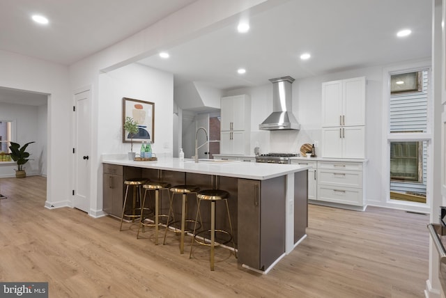 kitchen with light wood-type flooring, wall chimney range hood, white cabinets, a kitchen breakfast bar, and sink