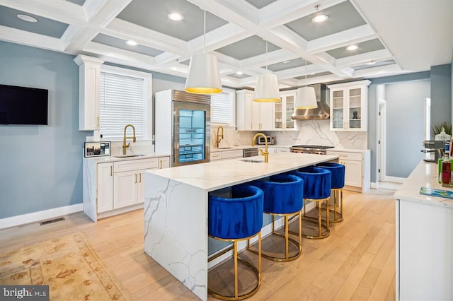 kitchen featuring white cabinets, wall chimney range hood, sink, a kitchen island with sink, and coffered ceiling