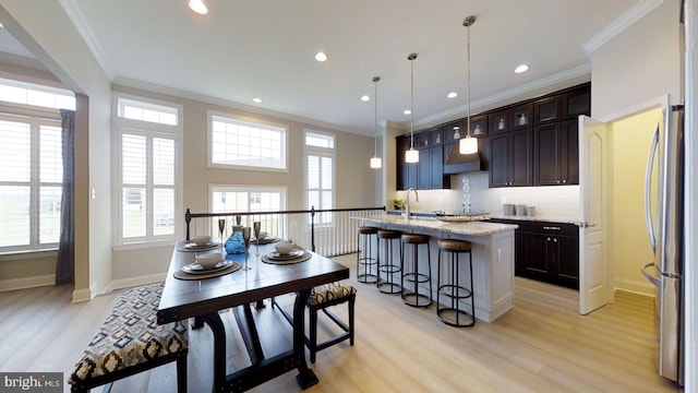 kitchen with light stone counters, a kitchen island with sink, decorative light fixtures, light hardwood / wood-style floors, and stainless steel refrigerator