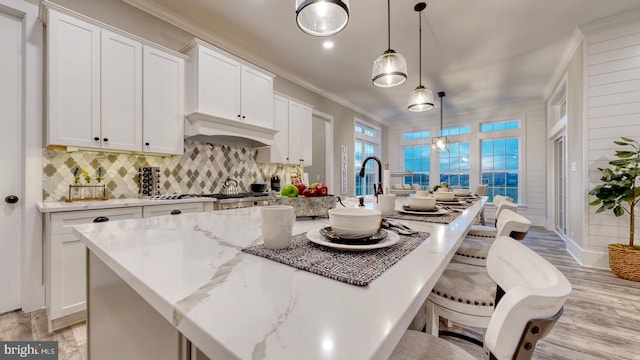 kitchen with hanging light fixtures, light hardwood / wood-style floors, an island with sink, white cabinets, and ornamental molding