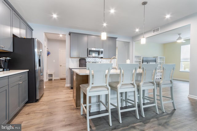 kitchen featuring stainless steel appliances, gray cabinetry, a kitchen island with sink, hanging light fixtures, and light hardwood / wood-style flooring