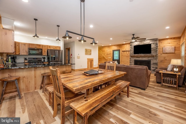 dining space with wood walls, a stone fireplace, sink, ceiling fan, and light hardwood / wood-style floors