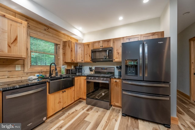 kitchen with appliances with stainless steel finishes, wooden walls, sink, dark stone countertops, and light hardwood / wood-style floors