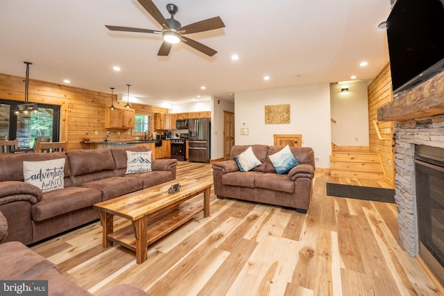 living room with light hardwood / wood-style floors, a stone fireplace, ceiling fan, and wooden walls