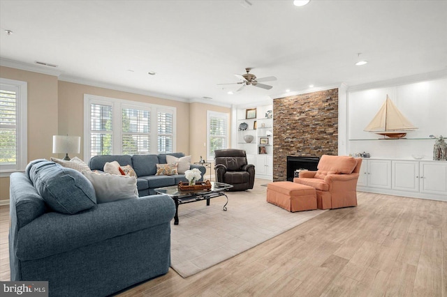 living room featuring built in features, light wood-type flooring, ceiling fan, a stone fireplace, and ornamental molding