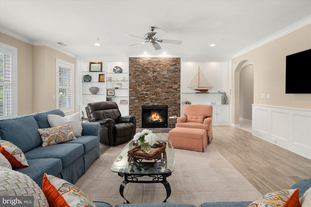 living room featuring built in shelves, crown molding, light wood-type flooring, a fireplace, and ceiling fan