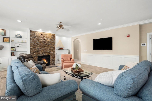 living room with light hardwood / wood-style floors, built in shelves, crown molding, ceiling fan, and a stone fireplace