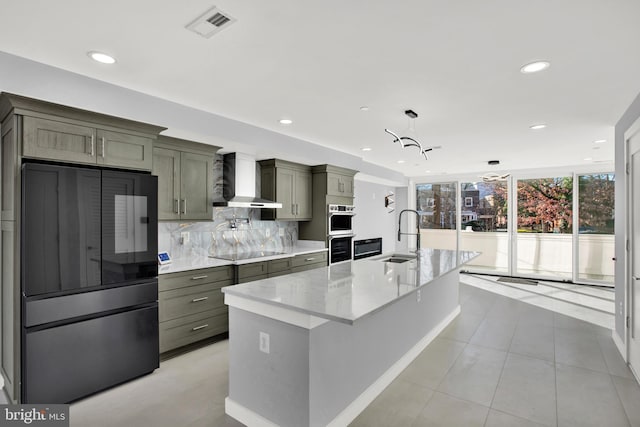 kitchen with wall chimney range hood, a sink, appliances with stainless steel finishes, backsplash, and an island with sink