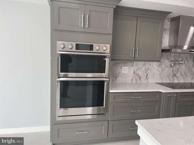 kitchen featuring wall chimney exhaust hood, stainless steel double oven, gray cabinetry, light stone countertops, and decorative backsplash