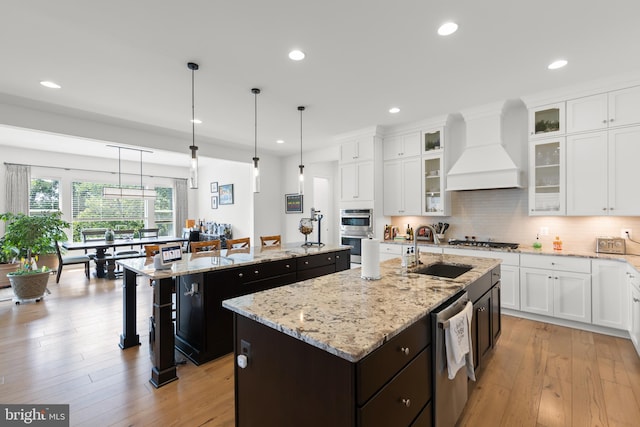 kitchen with light hardwood / wood-style flooring, custom exhaust hood, light stone counters, and a center island with sink