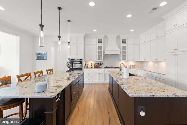 kitchen with backsplash, premium range hood, an island with sink, light hardwood / wood-style floors, and hanging light fixtures