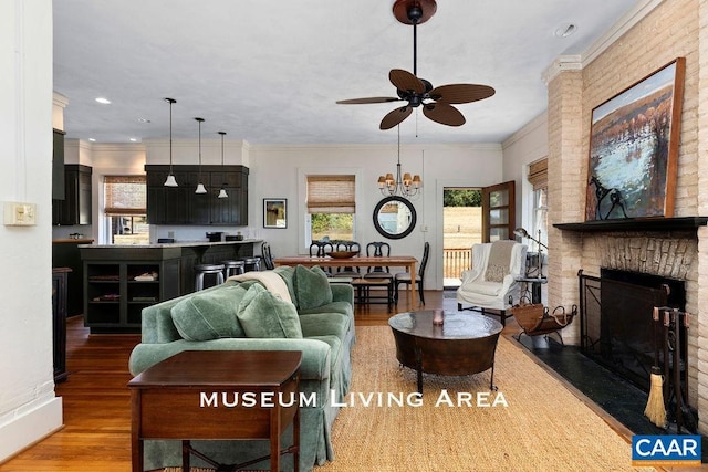 living room featuring hardwood / wood-style floors, ceiling fan with notable chandelier, a brick fireplace, and crown molding