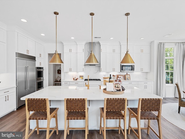 kitchen featuring stainless steel appliances, wall chimney range hood, decorative light fixtures, a kitchen island with sink, and a breakfast bar