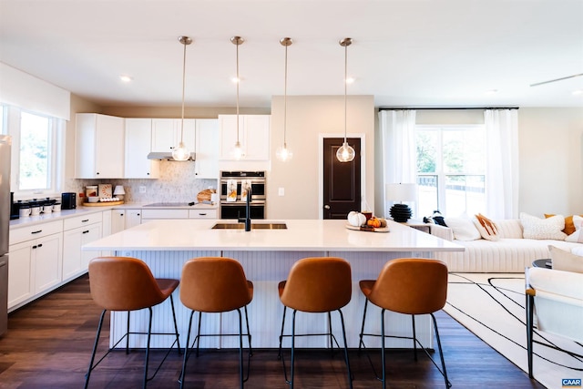 kitchen with double oven, a kitchen island with sink, sink, white cabinetry, and hanging light fixtures