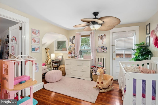 bedroom featuring wood-type flooring, radiator heating unit, a nursery area, and ceiling fan