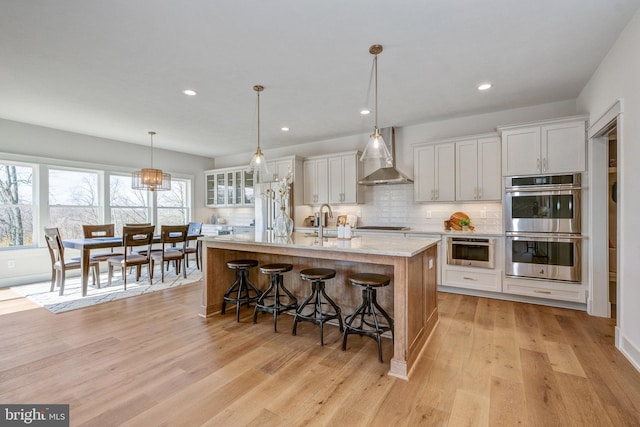 kitchen featuring light hardwood / wood-style flooring, white cabinets, a kitchen island with sink, wall chimney exhaust hood, and stainless steel double oven