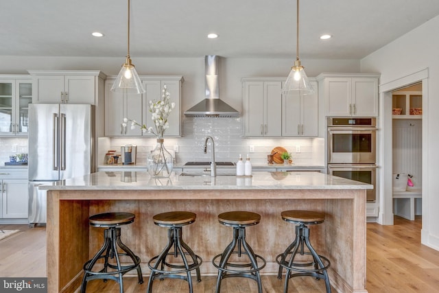 kitchen featuring light hardwood / wood-style flooring, backsplash, an island with sink, stainless steel appliances, and wall chimney exhaust hood