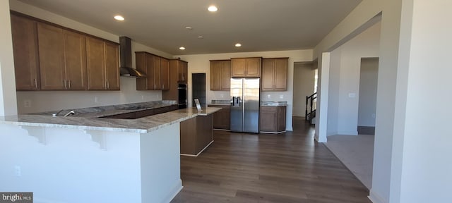 kitchen featuring light stone counters, kitchen peninsula, wall chimney range hood, stainless steel refrigerator with ice dispenser, and a breakfast bar area