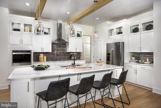 kitchen with appliances with stainless steel finishes, beamed ceiling, wall chimney range hood, hanging light fixtures, and backsplash