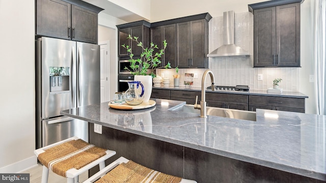 kitchen featuring stainless steel appliances, dark brown cabinets, decorative backsplash, dark stone countertops, and wall chimney exhaust hood