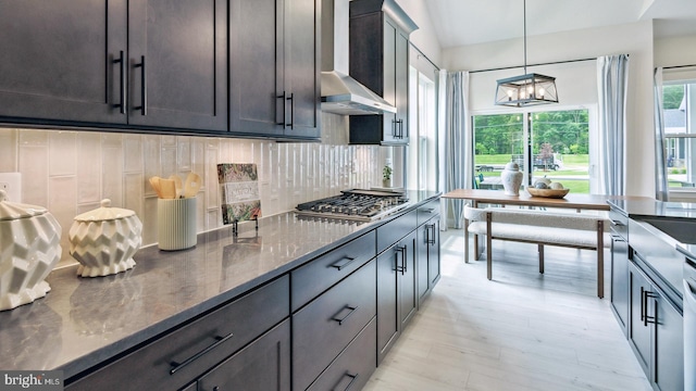 kitchen featuring wall chimney range hood, decorative backsplash, stainless steel gas stovetop, light wood-type flooring, and dark stone countertops