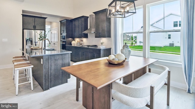 kitchen with light wood-type flooring, wall chimney exhaust hood, a center island with sink, appliances with stainless steel finishes, and backsplash