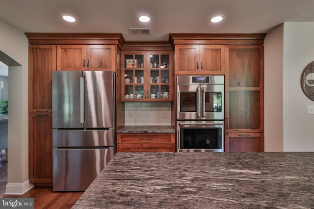 kitchen with dark wood-type flooring, backsplash, appliances with stainless steel finishes, and dark stone counters