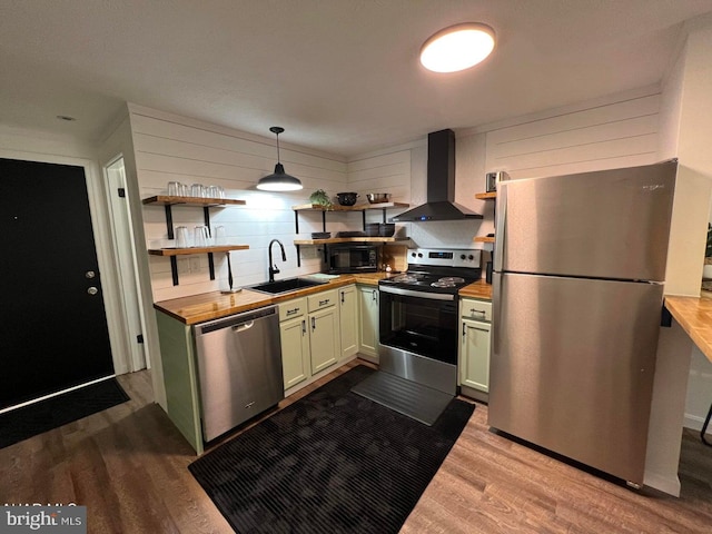 kitchen featuring wall chimney range hood, sink, hanging light fixtures, light wood-type flooring, and appliances with stainless steel finishes