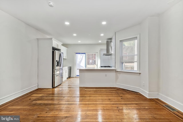 kitchen featuring wall chimney exhaust hood, kitchen peninsula, white cabinets, appliances with stainless steel finishes, and light hardwood / wood-style floors