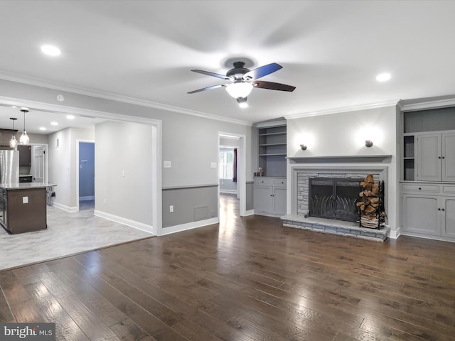 unfurnished living room with dark wood-type flooring, built in features, ceiling fan, ornamental molding, and a stone fireplace