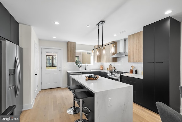 kitchen with pendant lighting, appliances with stainless steel finishes, wall chimney exhaust hood, a kitchen island, and light wood-type flooring