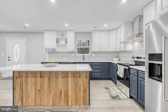 kitchen featuring white cabinetry, a center island, stainless steel appliances, blue cabinetry, and wall chimney range hood